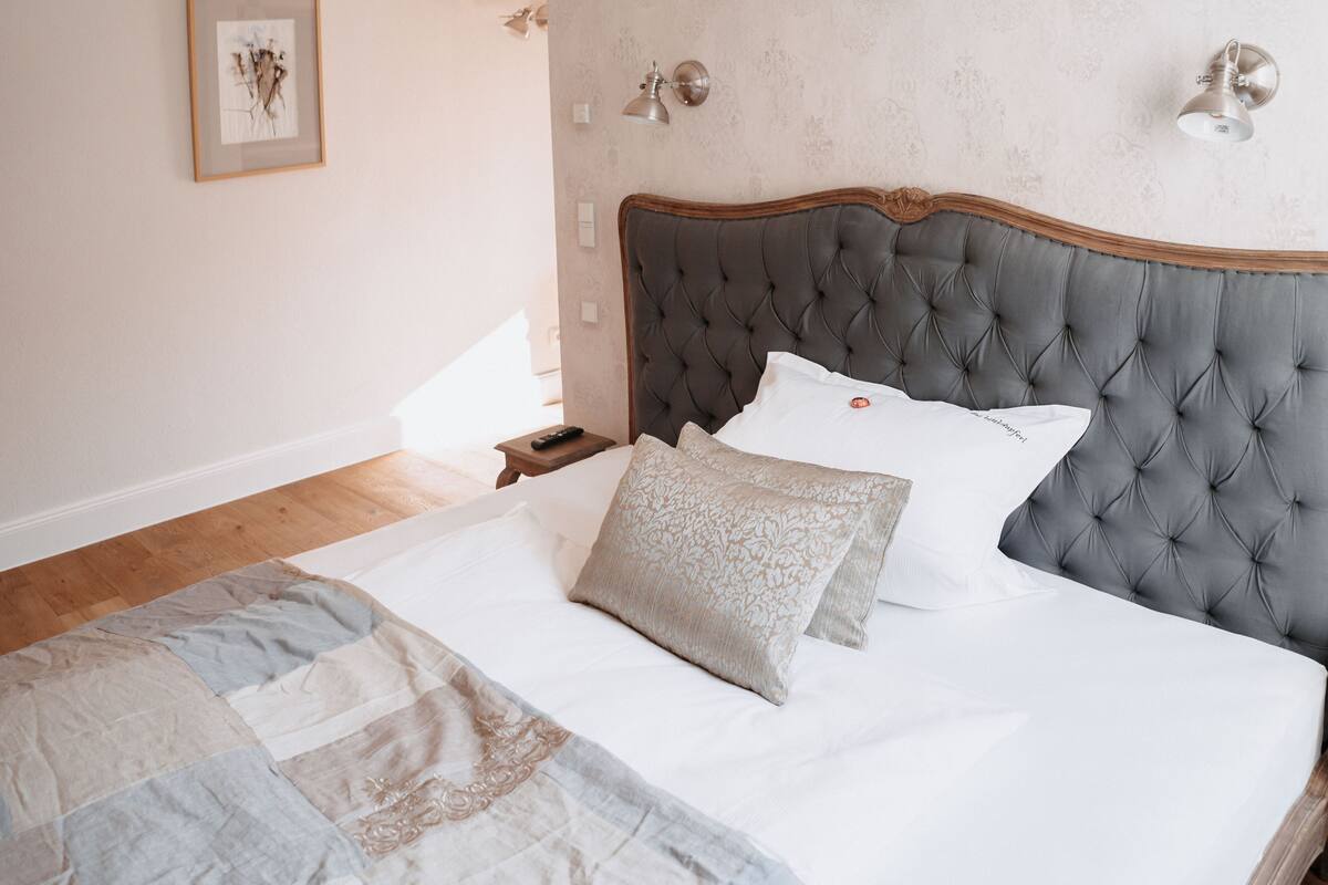 Een opgemaakt bed met een grijs bekleden hoofdeinde, witte lakens en een geborduurde plaid.