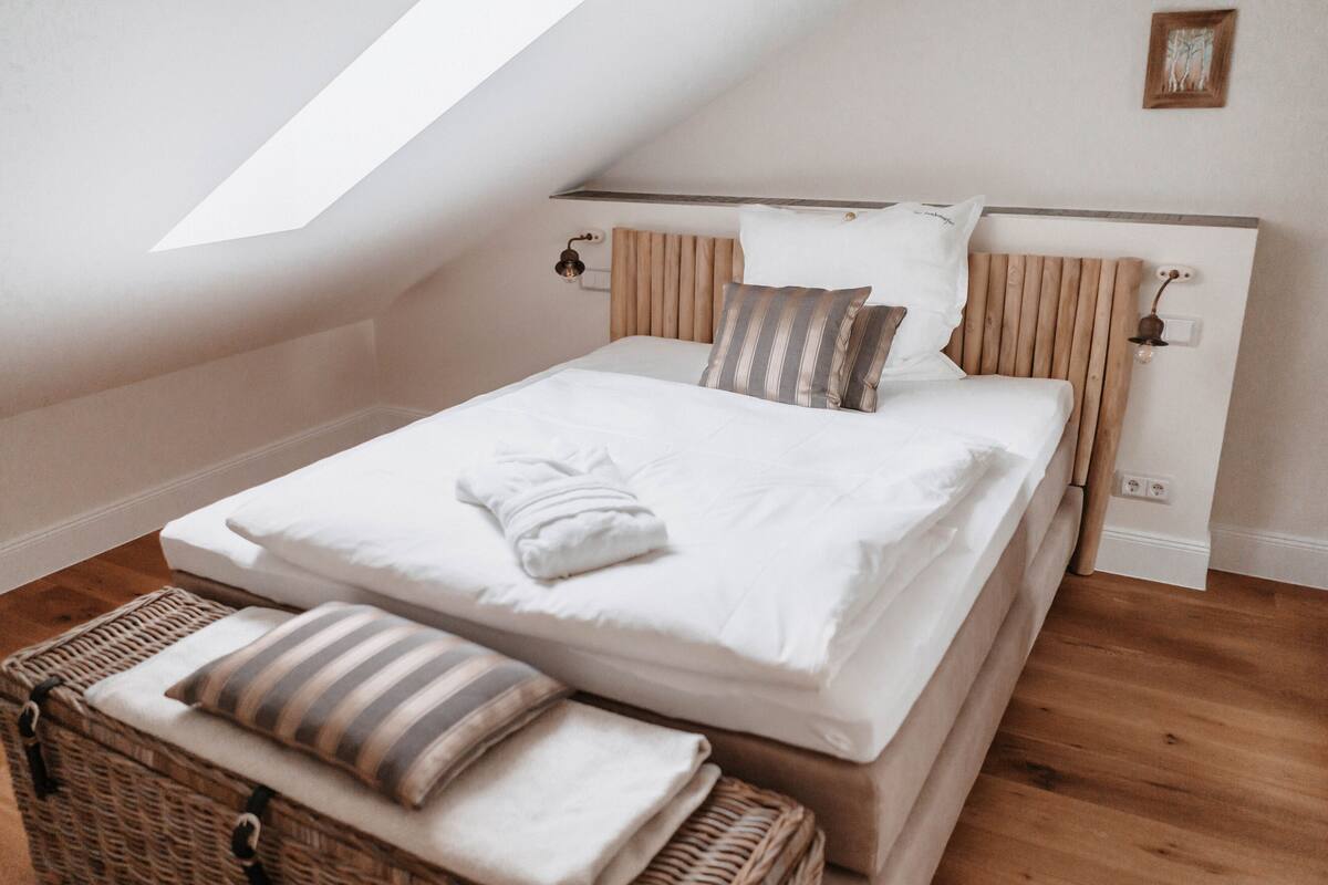 Een slaapkamer met een schuine kap, een houten hoofdbord, een bed met witte lakens, gestreepte kussens en een rieten mand met opgevouwen handdoeken.