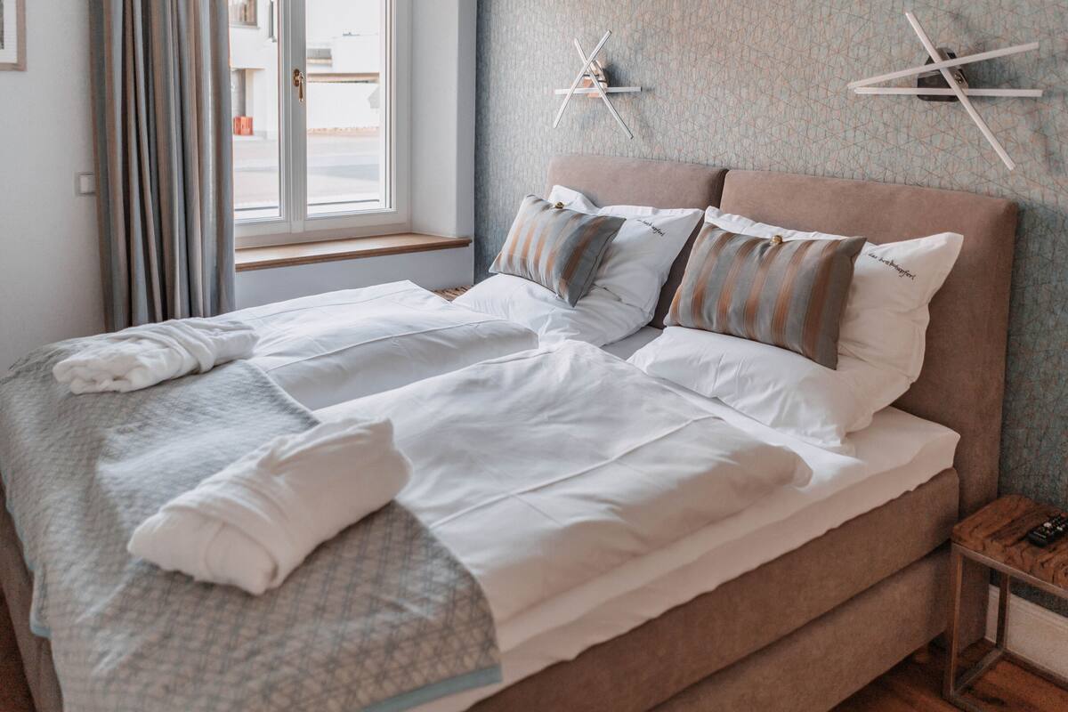 Een net opgemaakt bed met witte lakens en gestreepte kussens, een nachtkastje met een afstandsbediening en een raam met gordijnen.