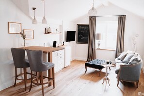 Una cocina moderna con una mesa de madera, dos taburetes de bar, un banco y un sofá. Hay una pizarra con texto, un televisor de pantalla plana y lámparas colgantes.