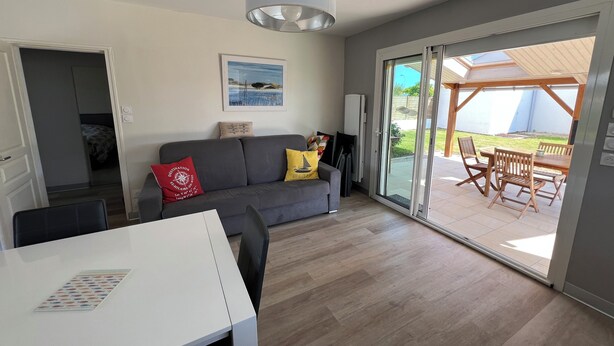 A modern living room with a grey sofa, a white table, and a sliding glass door leading to an outdoor patio with wooden furniture. - Comfortable vacation home ideally located between beach and town center (Saint-Hilaire-de-Riez)