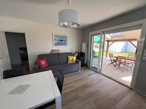 Een moderne woonkamer met een grijze bank, een witte tafel en een schuifdeur naar een terras buiten met houten meubels.