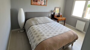 A bedroom with a bed, a bedside table, a lamp, a framed picture on the wall, and a window. - Comfortable vacation home ideally located between beach and town center (Saint-Hilaire-de-Riez)
