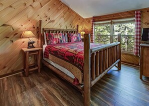 A wooden cabin bedroom with a bed, a nightstand, a lamp, and a window with a view of trees. - Bear Creek Ridge #2120 (Sevierville)