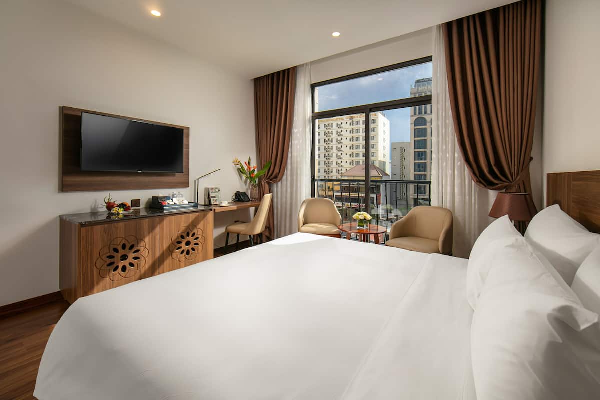 Ein Hotelzimmer mit einem großen Bett, einem Schreibtisch mit Flachbildfernseher, zwei Stühlen, einem kleinen Tisch mit Blumenvase und Blick auf eine Stadtlandschaft durch große Fenster.