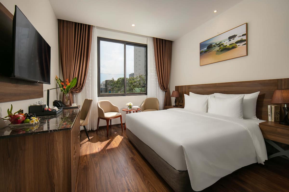 Ein Hotelzimmer mit einem großen Bett, einem an der Wand montierten Fernseher, einem Schreibtisch mit Blumen und einem Vorhangfenster.