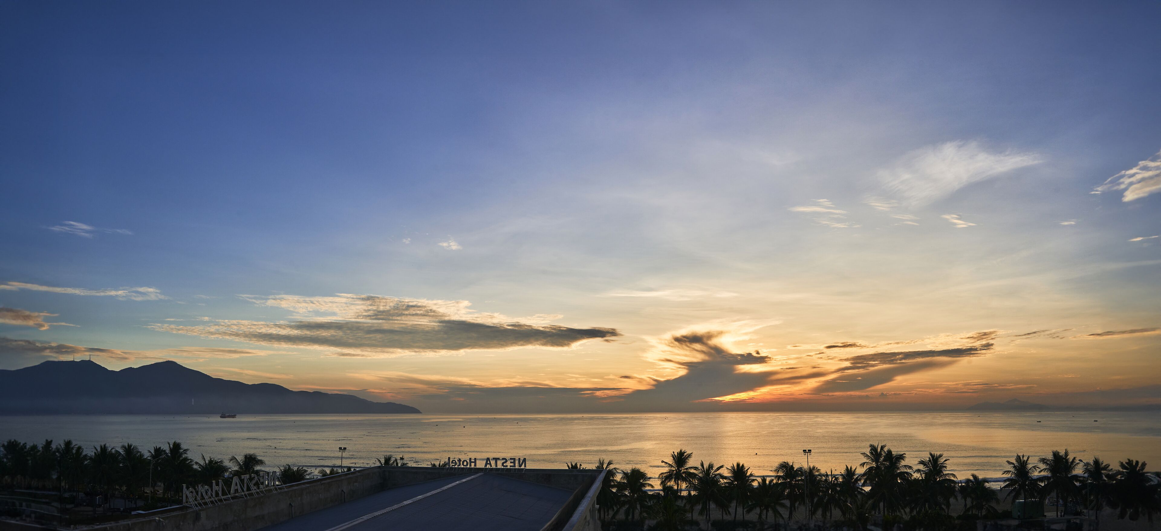 A coastal view with a sunset, palm trees, and a distant mountain range.