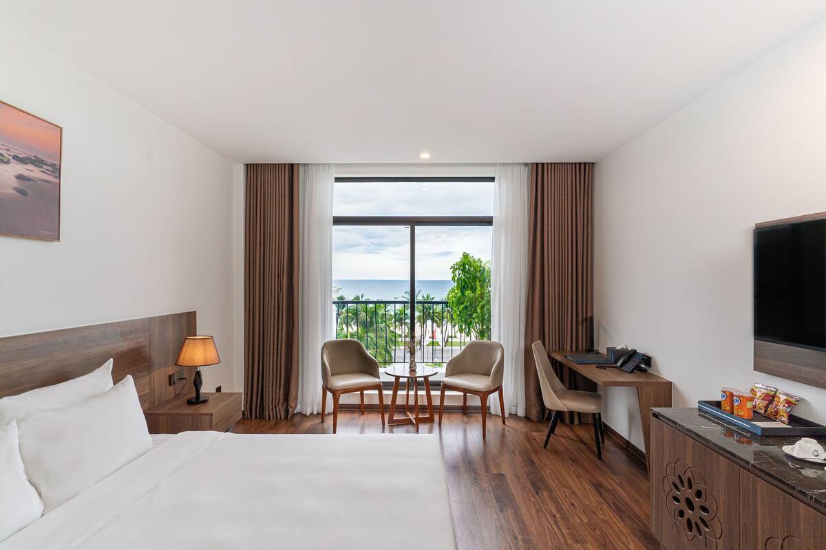 Ein Hotelzimmer mit Bett, Schreibtisch, zwei Stühlen, Fernseher und einem Balkon mit Meerblick.