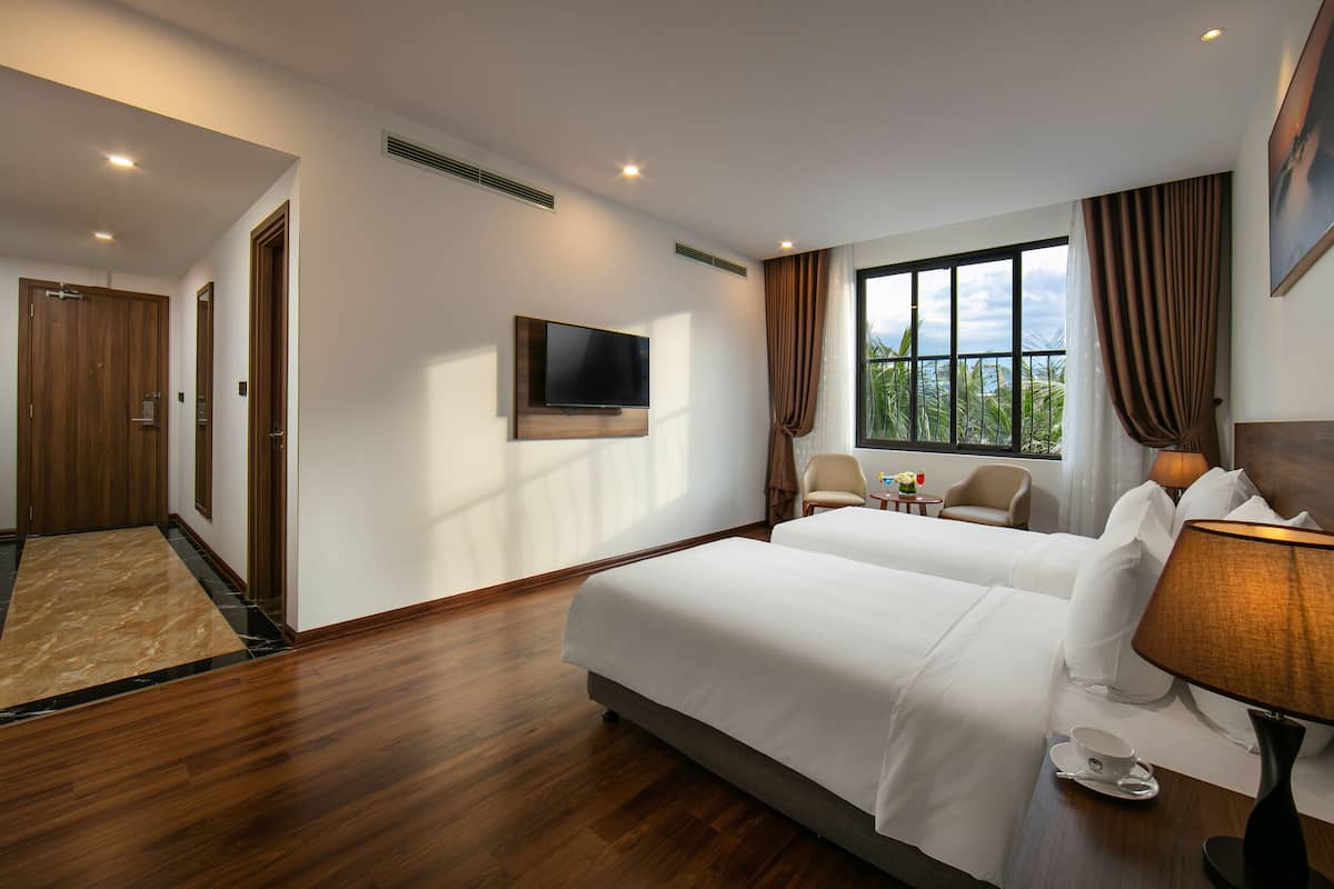 Ein Hotelzimmer mit zwei Betten, einem Fernseher, einem Fenster mit Vorhängen, einer Lampe und einem kleinen Tisch mit einer Tasse.