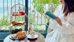 Una persona leyendo un libro en una mesa con pasteles y café, con vista a un balcón que da al mar.