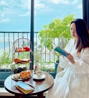 A person reading a book at a table with pastries and coffee, overlooking a balcony with a view of the sea.
