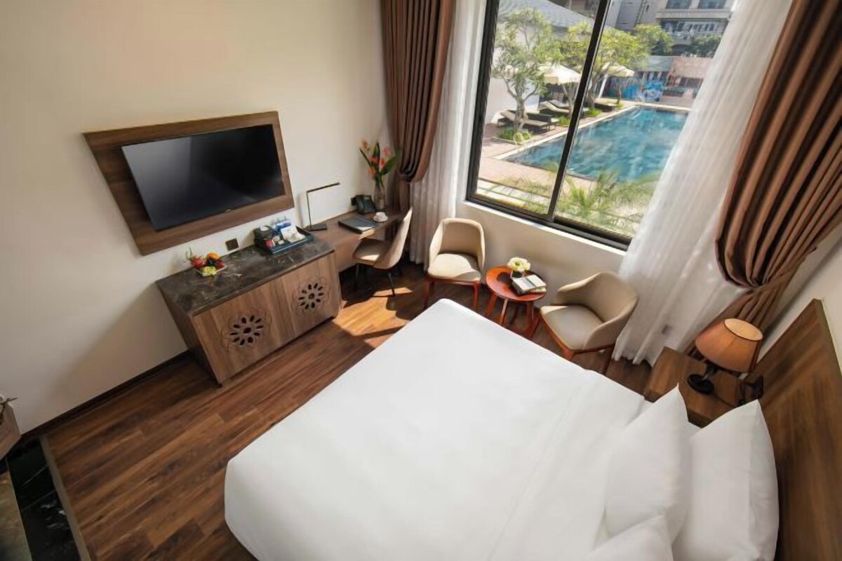 A hotel room with a bed, a TV mounted on the wall, a desk with a chair, a small table with a lamp, and a view of a swimming pool through large windows.