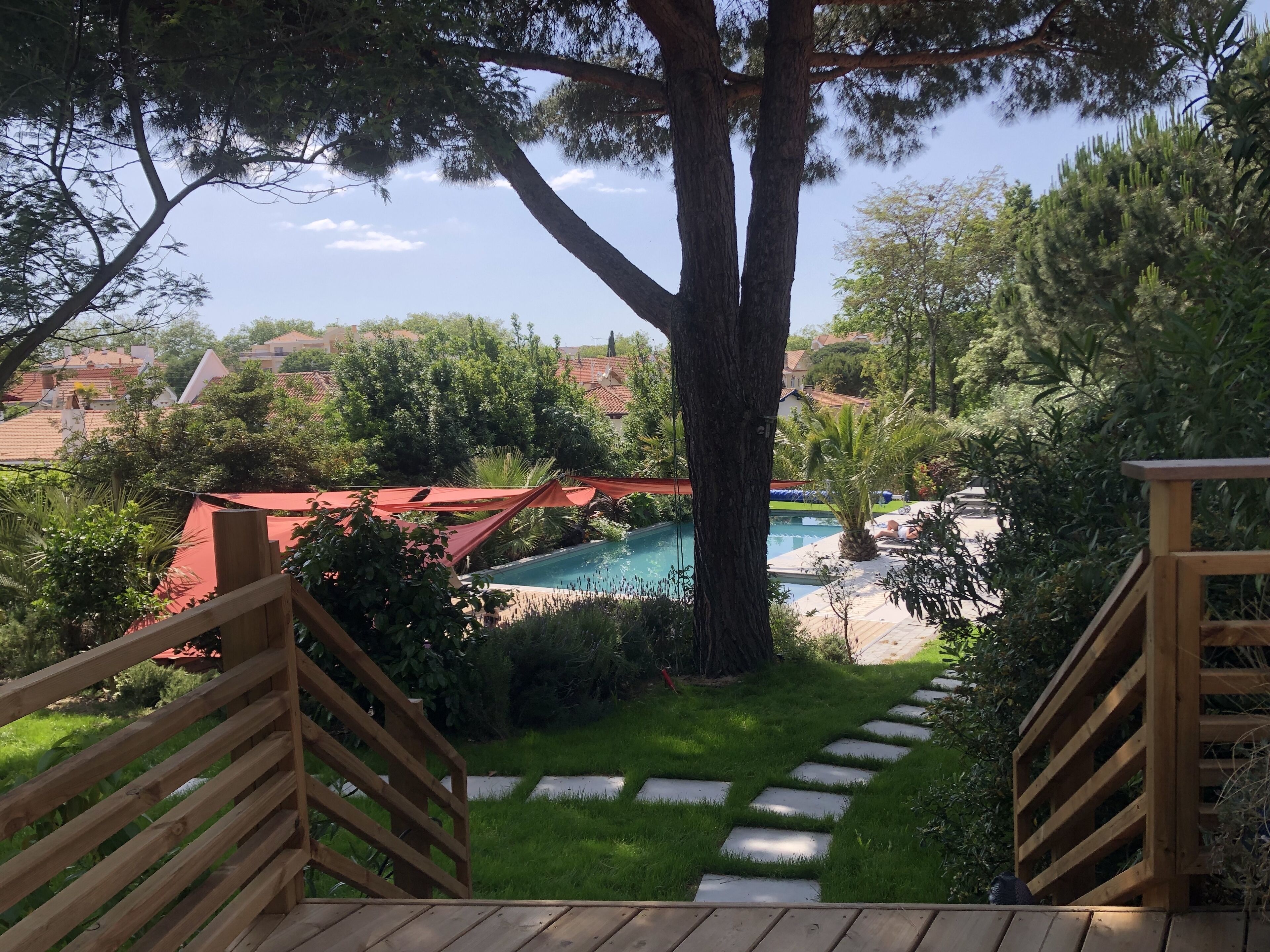 Un escalier en bois mène à un espace piscine entouré de verdure, avec en arrière-plan une vue sur des maisons.