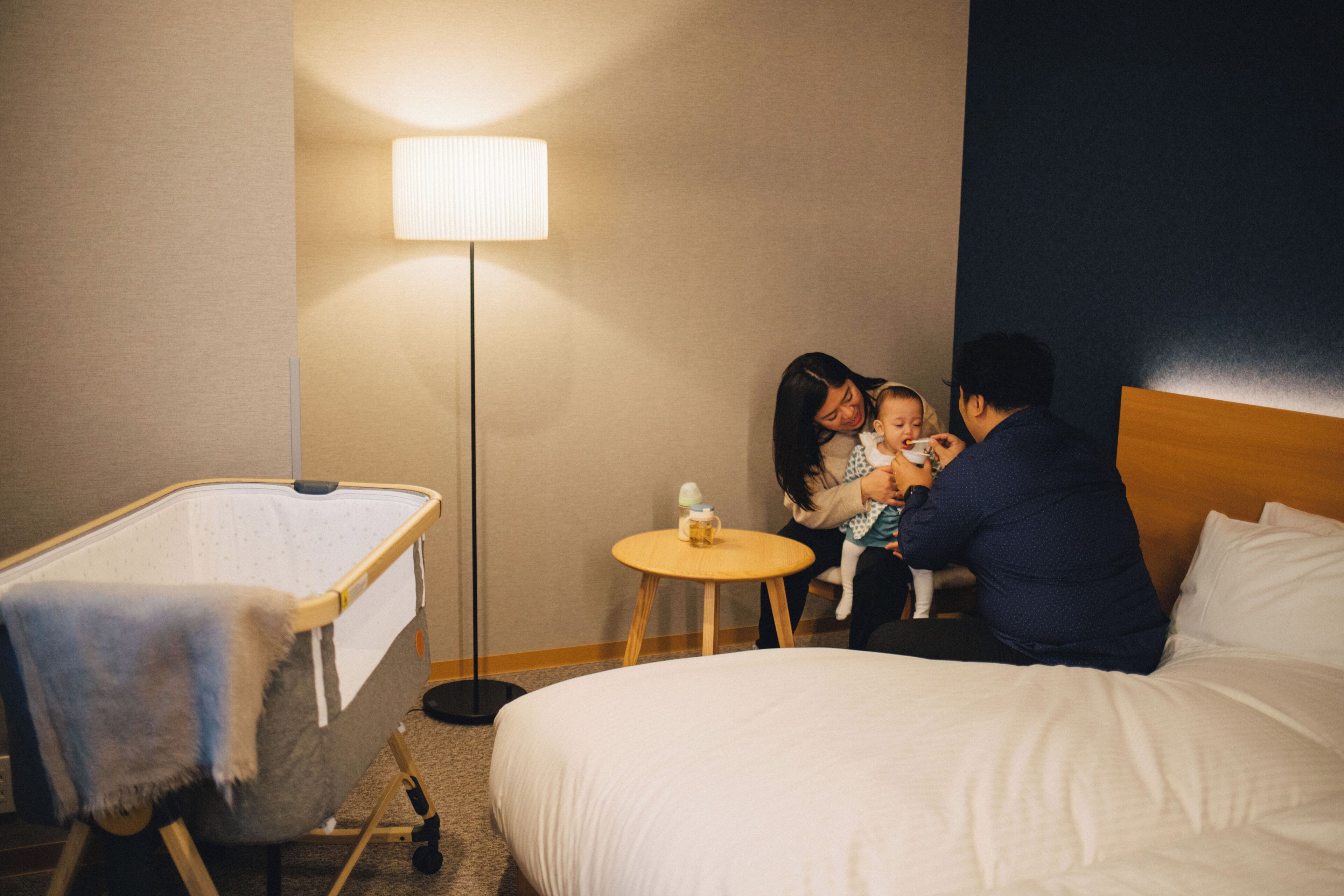 Een hotelkamer met een reisbedje, een nachtkastje met een lamp en een bed.
