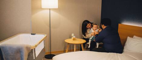 A hotel room with a crib, a bedside table with a lamp, and a bed.