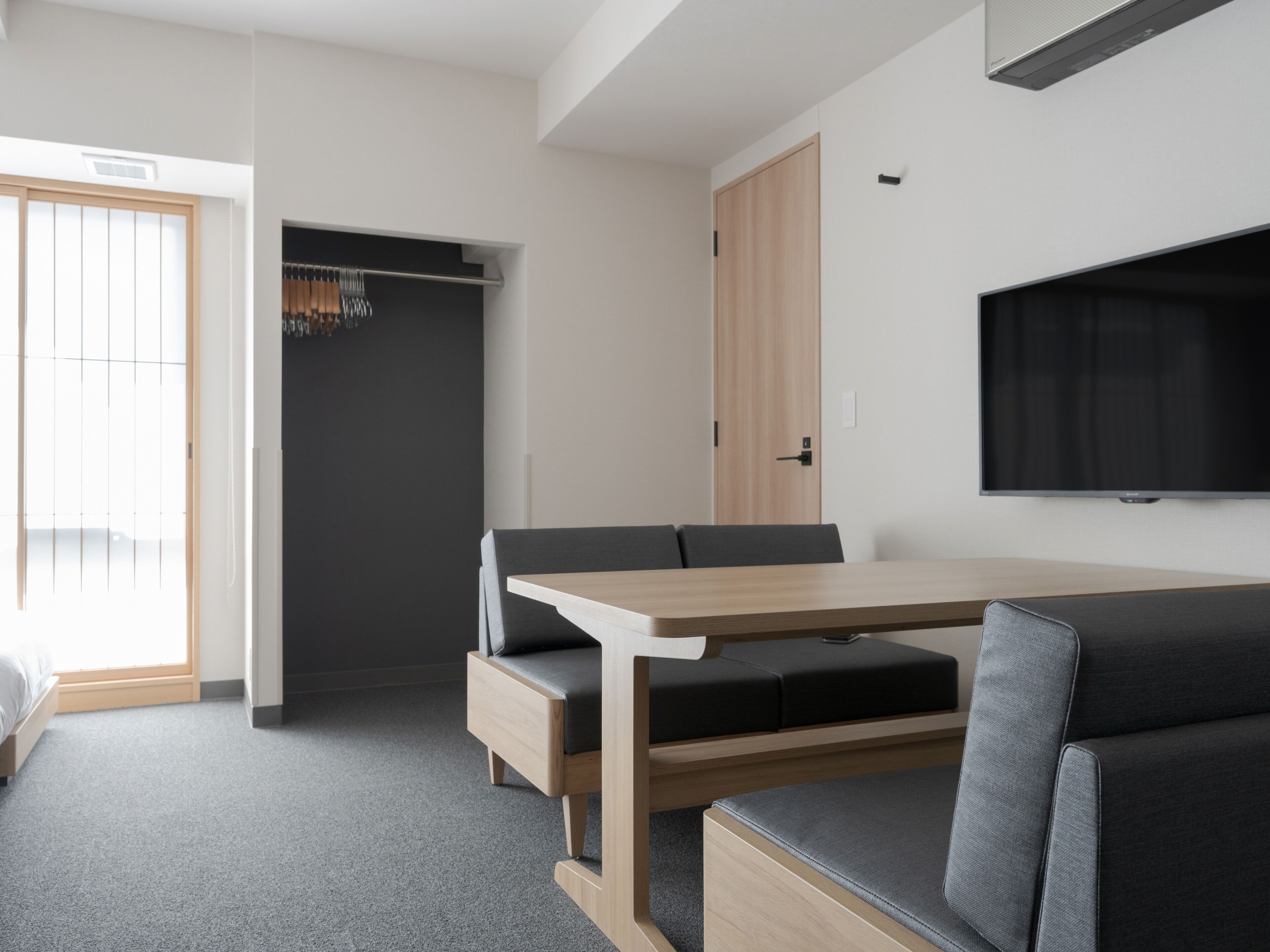 A modern hotel room with a wooden dining table, grey sofa, and a TV mounted on the wall.
