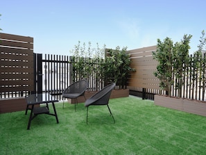 Um pátio externo com duas cadeiras de vime e uma mesa de vidro, cercado por um muro de madeira e vegetação.