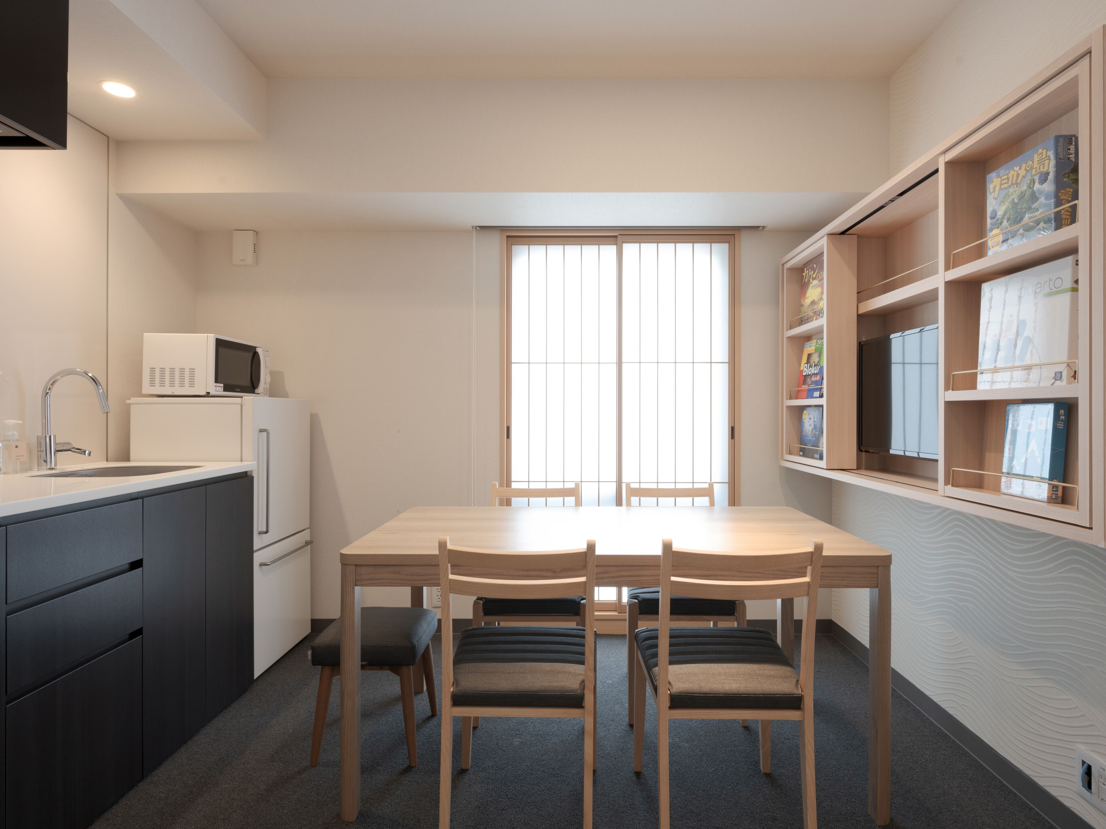 Una cocina compacta con microondas, fregadero y armarios. Hay una mesa de comedor con sillas y una ventana con persianas verticales.