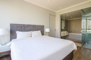 Quarto moderno com uma cama grande, luminárias de cabeceira, banheiro com paredes de vidro e vista para a cidade.