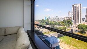 Ein Zimmer mit großem Fenster und Blick auf eine Stadtlandschaft, ein beiges Sofa und ein kleiner Tisch mit einem Buch.