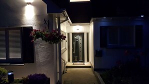 A modern house with a dark entrance door, a hanging flower basket, and a light fixture above the door. - Beautifully  Presented One Bedroom Romantic Home & Private Hot Tub. (WEST LOOE)
