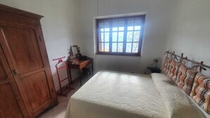 A bedroom with a bed, a wooden wardrobe, a desk with a chair, a window, and a wall-mounted shelf.