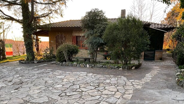 A stone-paved courtyard with a rustic house, surrounded by greenery and a stone wall.