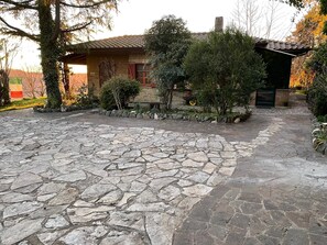 Un pati empedrat de pedra amb una casa rústica, envoltat de vegetació i una paret de pedra.