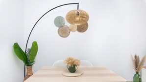 Un comedor con una mesa de madera, sillas blancas, una lámpara de pie tejida y un jarrón verde con plumas de plumas.