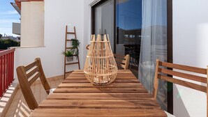 Una mesa de madera con sillas en un balcón, una silla de mimbre y una escalera apoyada en la pared.
