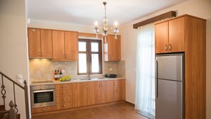 A kitchen with wooden cabinets, a stainless steel refrigerator, a chandelier, and a window with curtains. - FLORENA VILLAS (Lefkada)