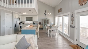 A spacious living area with a high ceiling, a dining table with chairs, a kitchen with modern appliances, and a balcony with a view.