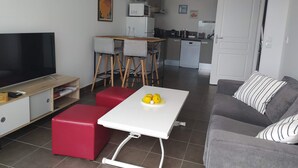 Een compacte woonruimte met een televisie, een eettafel met stoelen, een keukenhoek en een bank.