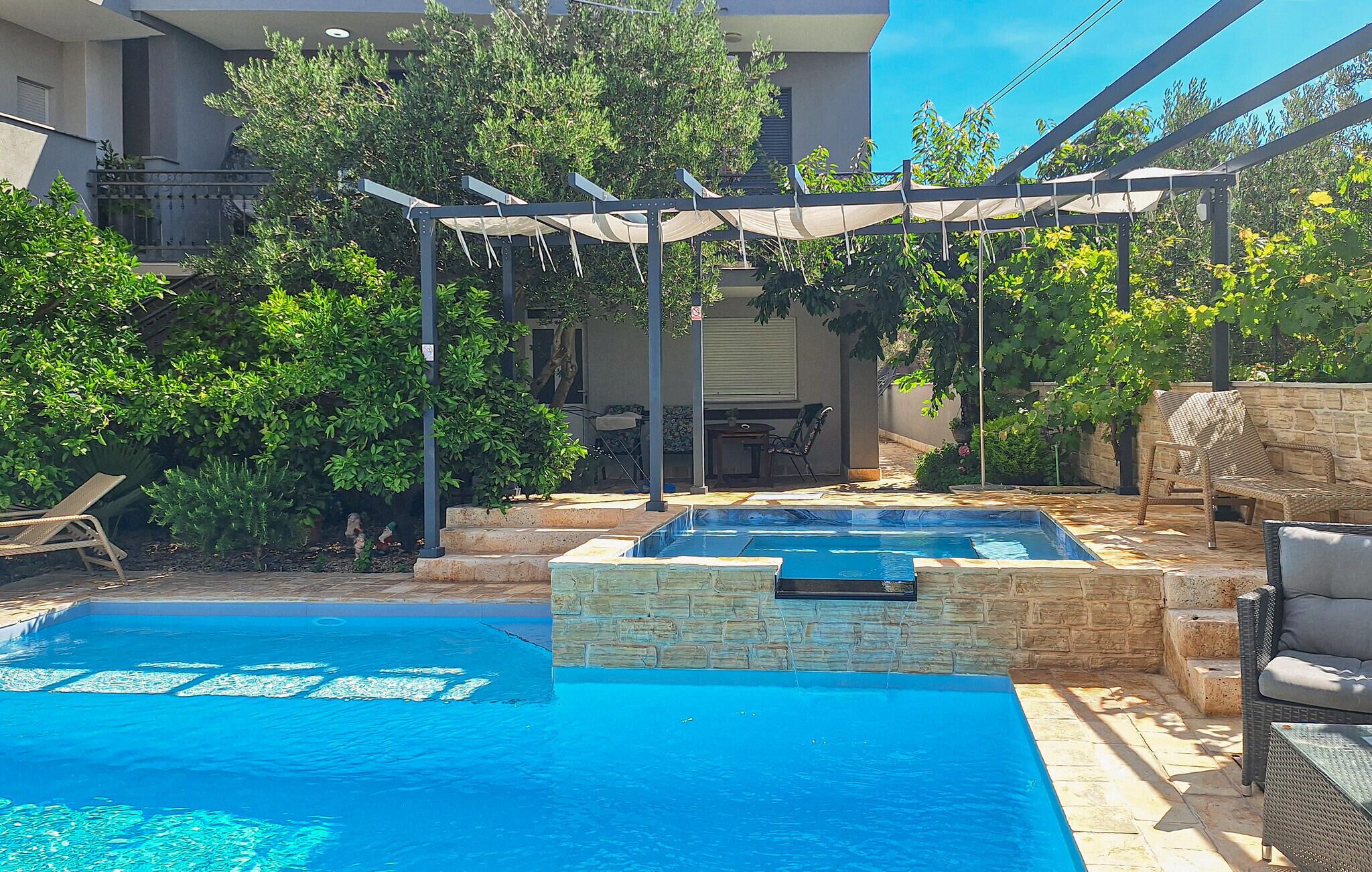 Una piscina con bordatura in pietra, un pergolato con tettoia e mobili da esterno.