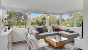 Ein sonnendurchflutetes Zimmer mit grauem Sofa, einem Glastisch und Blick auf einen Garten durch große Fenster.