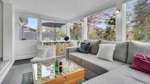Ein geräumiges Wohnzimmer mit grauem Sofa, einem Glastisch und großen Fenstern mit Blick auf Bäume und einen Zaun.