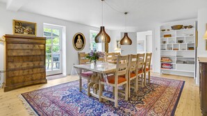 Un comedor con una mesa de madera, sillas, un tapete grande, luces colgantes y una estantería.