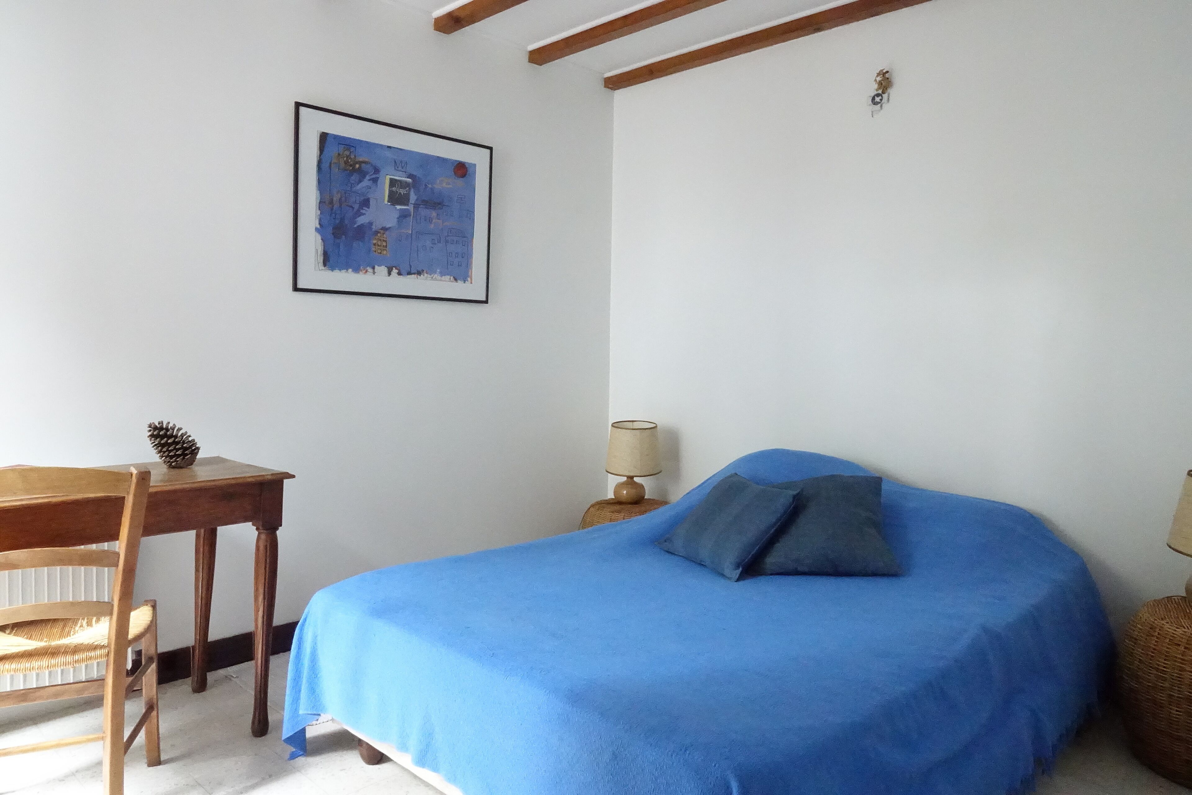 Un dormitorio con una cama azul, una silla de madera, una mesita, una lámpara y un cuadro en la pared.