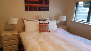 A neatly made bed with white bedding and a decorative pillow, two bedside lamps, a small drawer unit, and a painting above the bed.