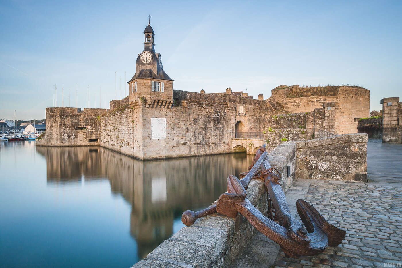 Un bâtiment historique en pierre, doté d’une tour horloge, d’un fossé et d’un quai en bois avec une ancre.