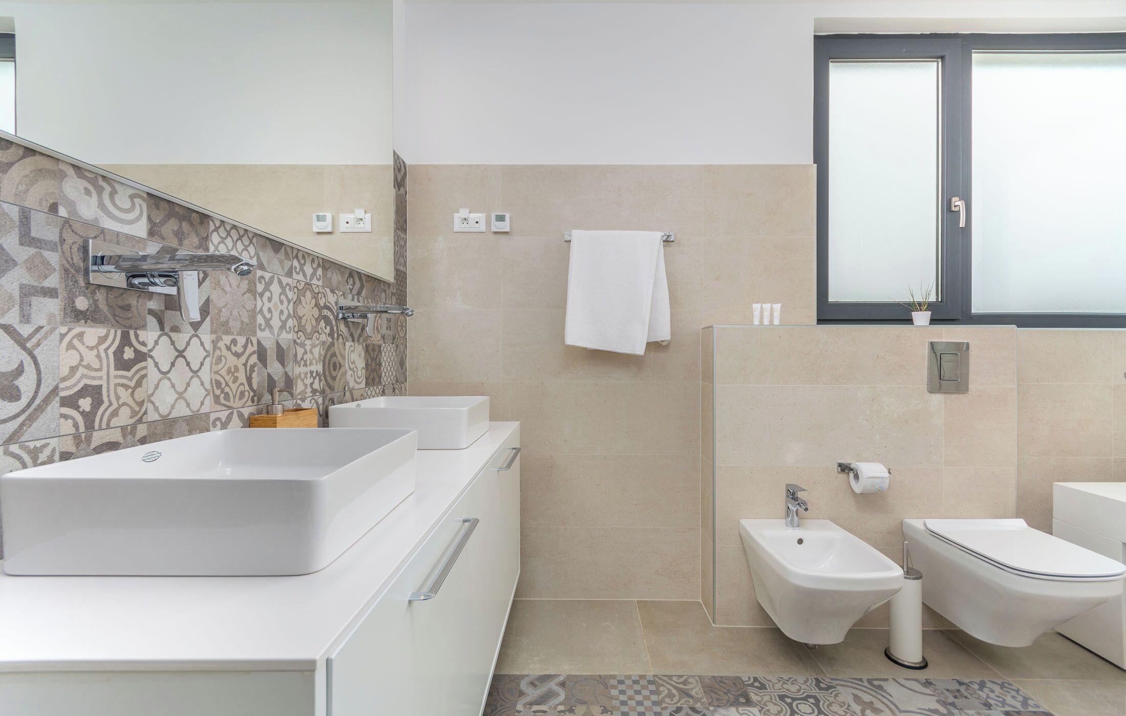 A modern bathroom with a large sink, bidet, and toilet, tiled walls, and a window with frosted glass.