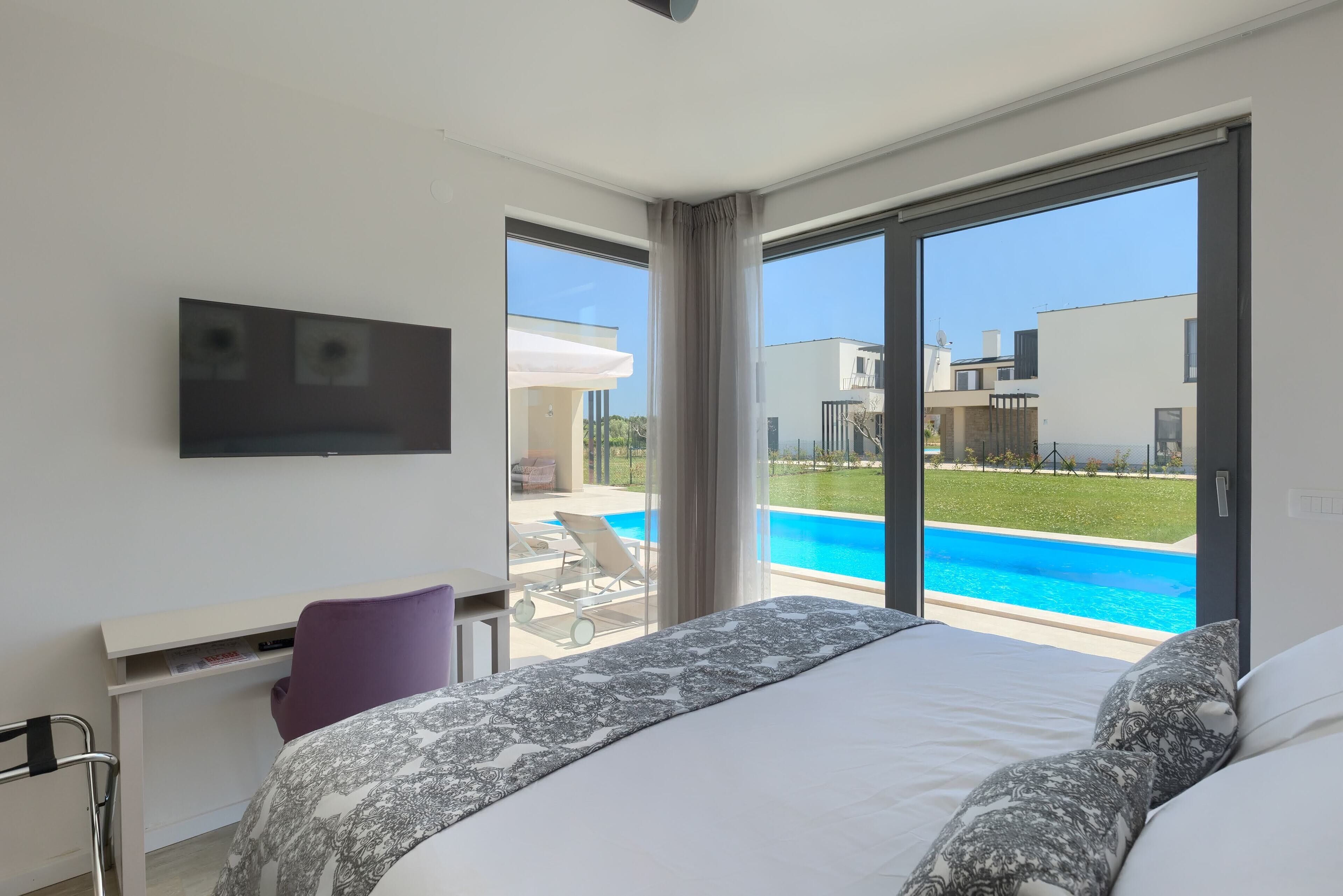 A bedroom with a large bed, a desk, a chair, and a TV mounted on the wall. It features a large window with a view of a swimming pool and a building outside.