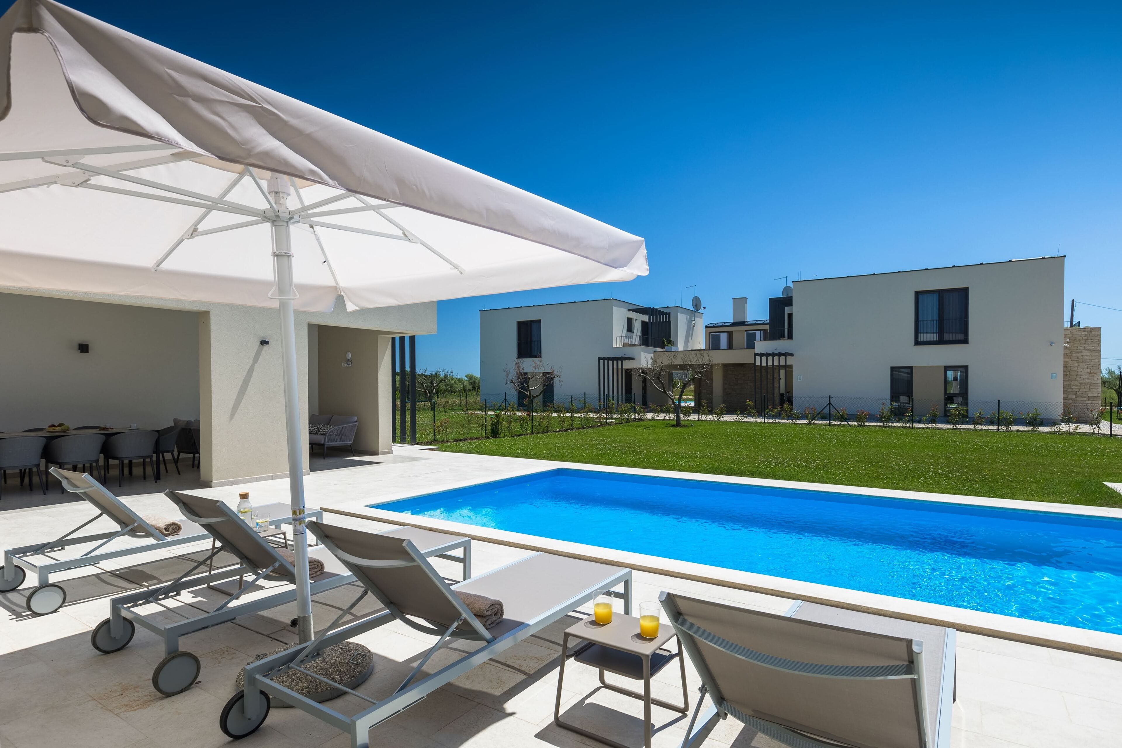 Zona de piscina moderna al aire libre con sillones de descanso y una mesa, rodeada de un césped cuidado y un edificio contemporáneo al fondo.