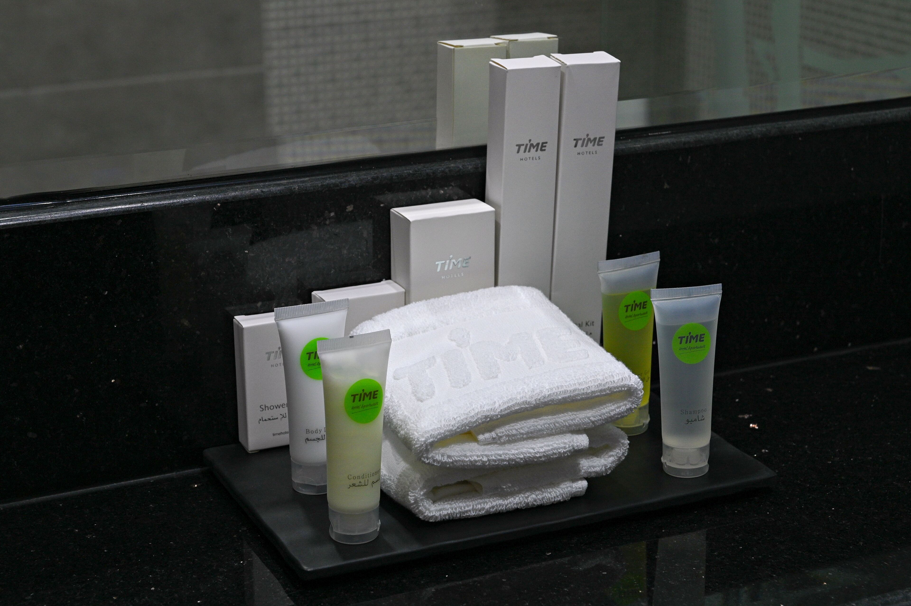 A hotel bathroom with a black countertop displaying toiletries, including a white towel, a green bottle, and a white box labeled "TIME HOTELS".
