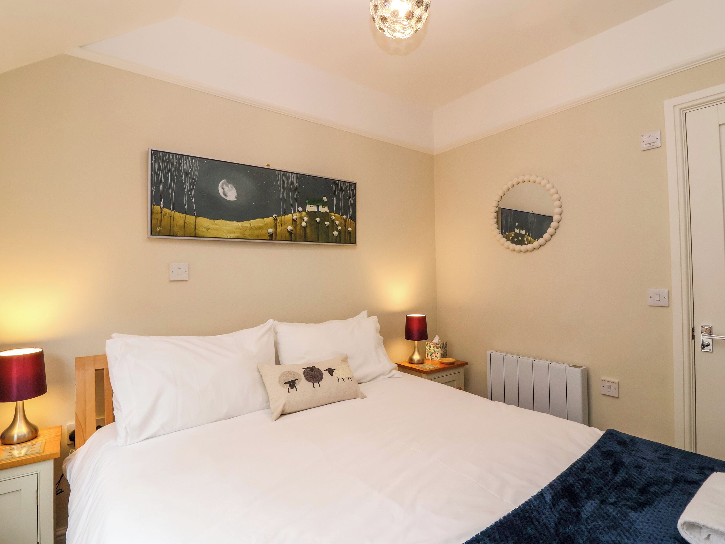 Un lit bien fait, avec du linge de lit blanc, un seul coussin décoratif et un plaid bleu. Au-dessus du lit, on peut voir un tableau représentant un paysage éclairé par la lune.