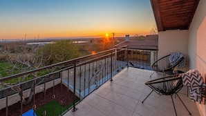 En balkon med udsigt til solnedgang, klinkegulv og to sorte stole med mønstrede hynder.