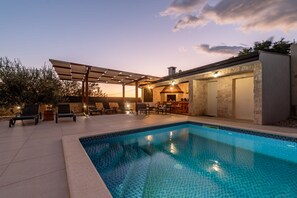 Uma piscina com pergolado e espreguiçadeiras, um edifício de pedra com porta e uma área de pátio com bar.
