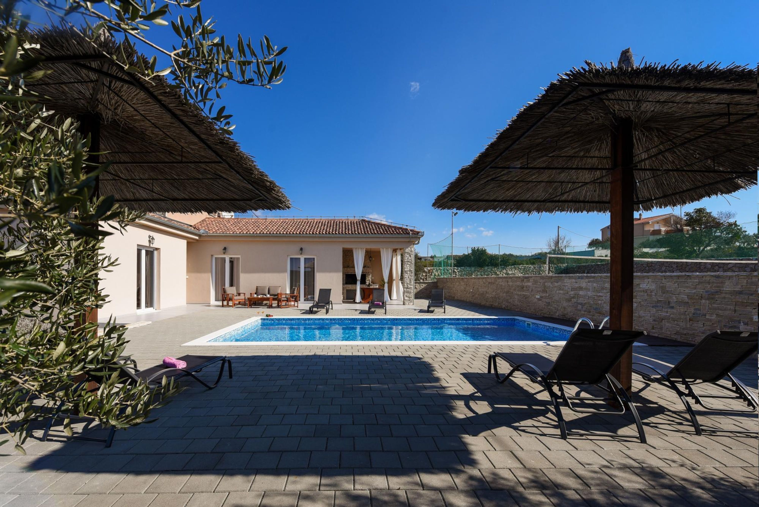Area piscina con sedie a sdraio e ombrelloni, un edificio con tetto in tegole, e un cielo azzurro limpido.