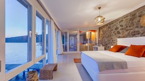 A modern bedroom with a large bed, a stone accent wall, and a view of the sea.