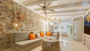 A modern living room with a stone wall, a gray sofa with orange cushions, a wooden coffee table, and a ceiling fan.